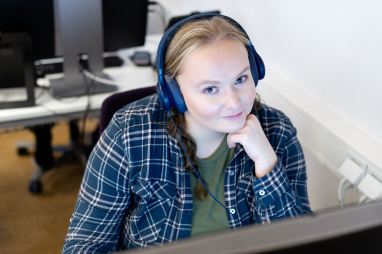 studerande ung kvinna på YH-utbildningen medicinsk vårdadministratör sitter framför dator med hörlurar på huvudet, och tittar leende in i kameran.