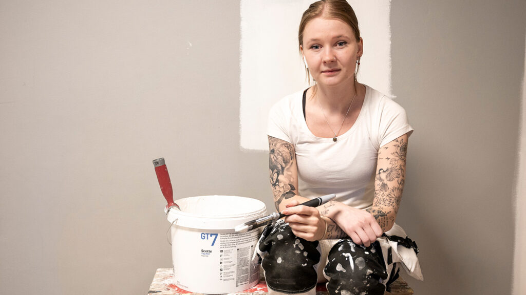 Kvinnlig studerande till målare. Sitter på en arbetsbänk i arbetskläder med penslar och en färghink.