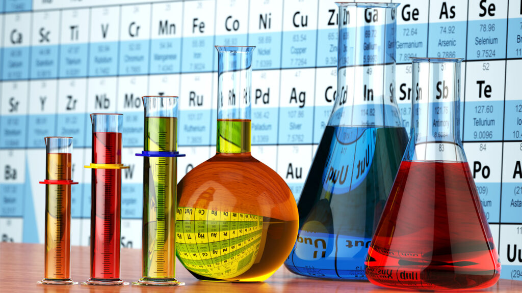 Bild med labb- och kemiutrustning. Kemiska glasvaror fyllda med färgglada vätskor. I bakgrunden tavla med kemiska formler.