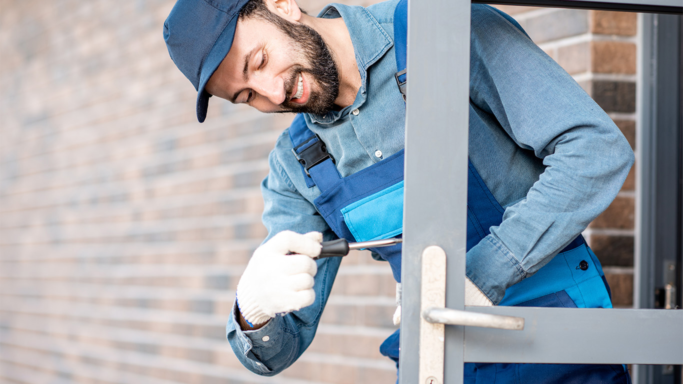 Bild. man i blåkläder och keps. Monterar lås i en dörr.
